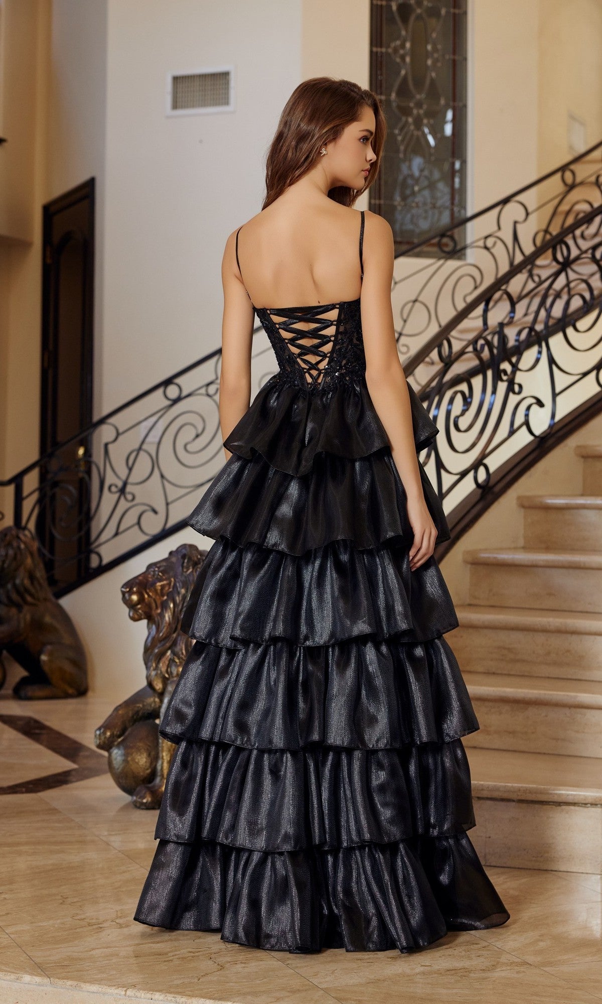 A woman stands indoors on a marble floor, facing away, wearing the Nox Anabel N1762 long prom dress with a ruffle tiered organza skirt and lace-up back. A staircase with ornate railing is seen in the background.